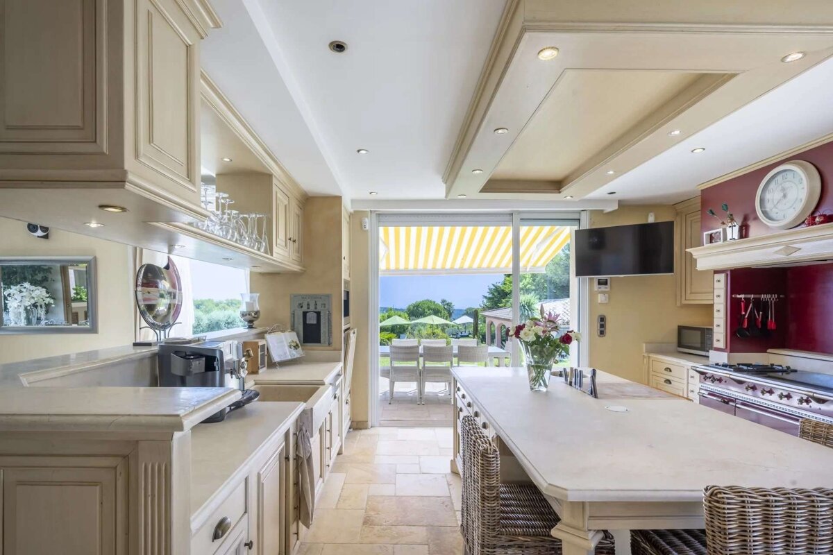 Elegant kitchen with cream cabinets, a large island, red accent wall, and sliding glass doors opening to a beautiful outdoor patio and scenic view.