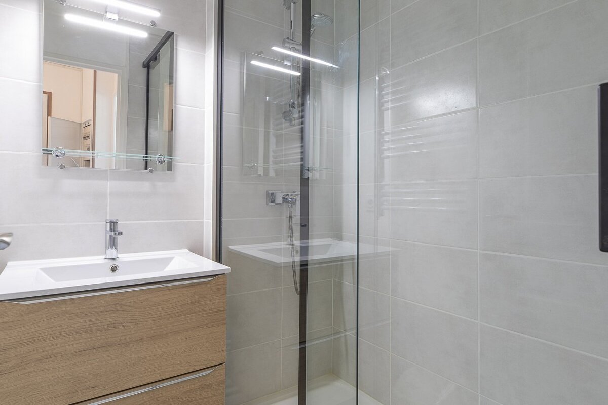 Bright modern bathroom with a light wood vanity, white sink, illuminated mirror, and a glass shower enclosure with grey tiles and a towel radiator.