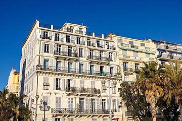 Elegant cream buildings with many windows and balconies line a street under a bright blue sky, alongside green palm trees.