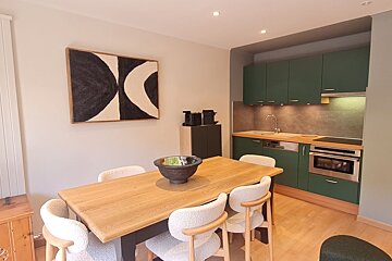 A dining area with a wooden table, white chairs, and abstract art next to a kitchen featuring green cabinets.