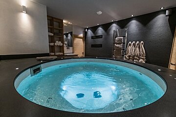 A modern indoor spa room with a round, glowing blue hot tub, dark textured walls, spotlights, wooden shelving with towels, and bathrobes.