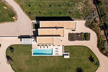Aerial view of a modern villa with three terracotta-roofed sections, a rectangular pool, manicured lawn, and gravel pathways amidst green landscape.