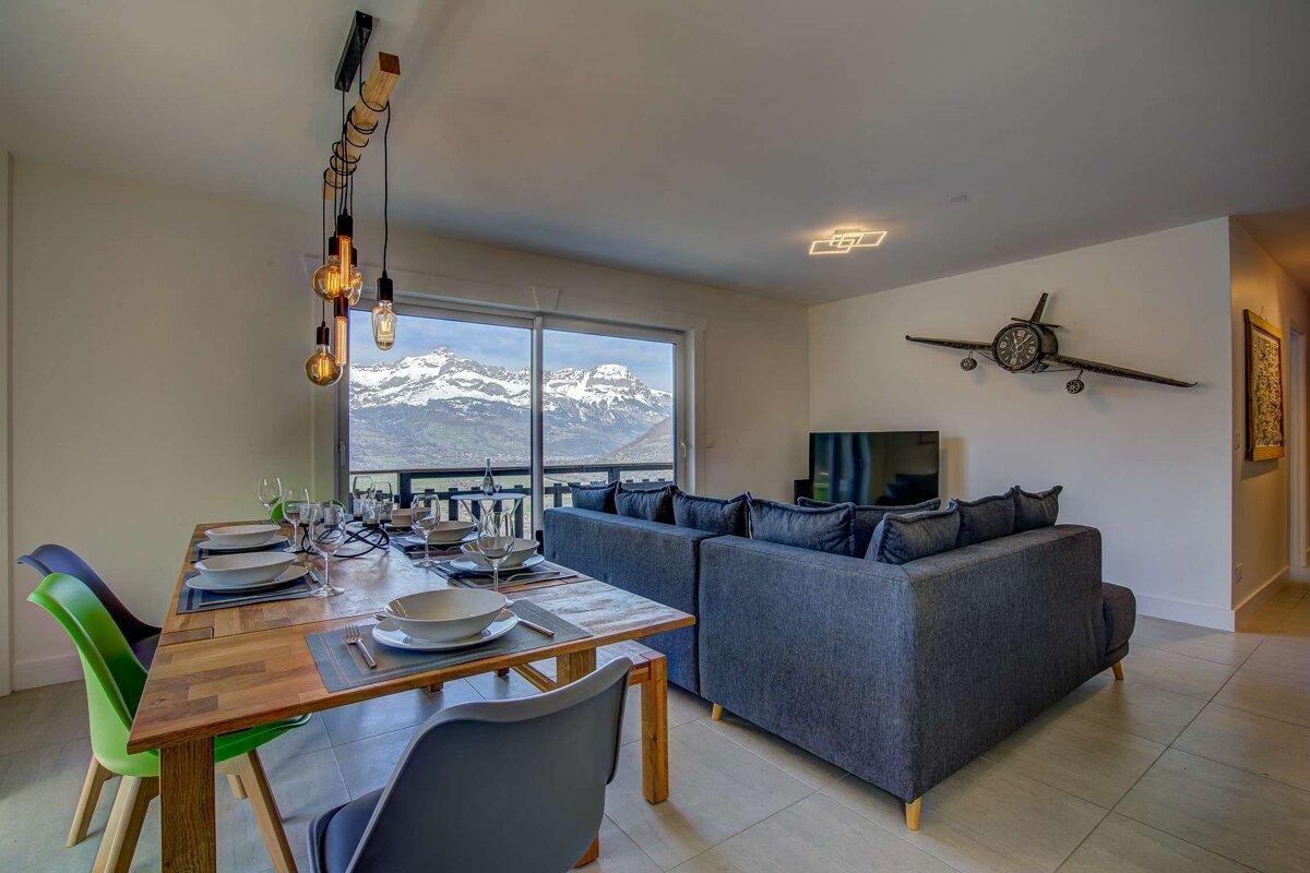 A modern living area featuring a wooden dining table and grey sofa, with a large window showing stunning snow-capped mountain views.
