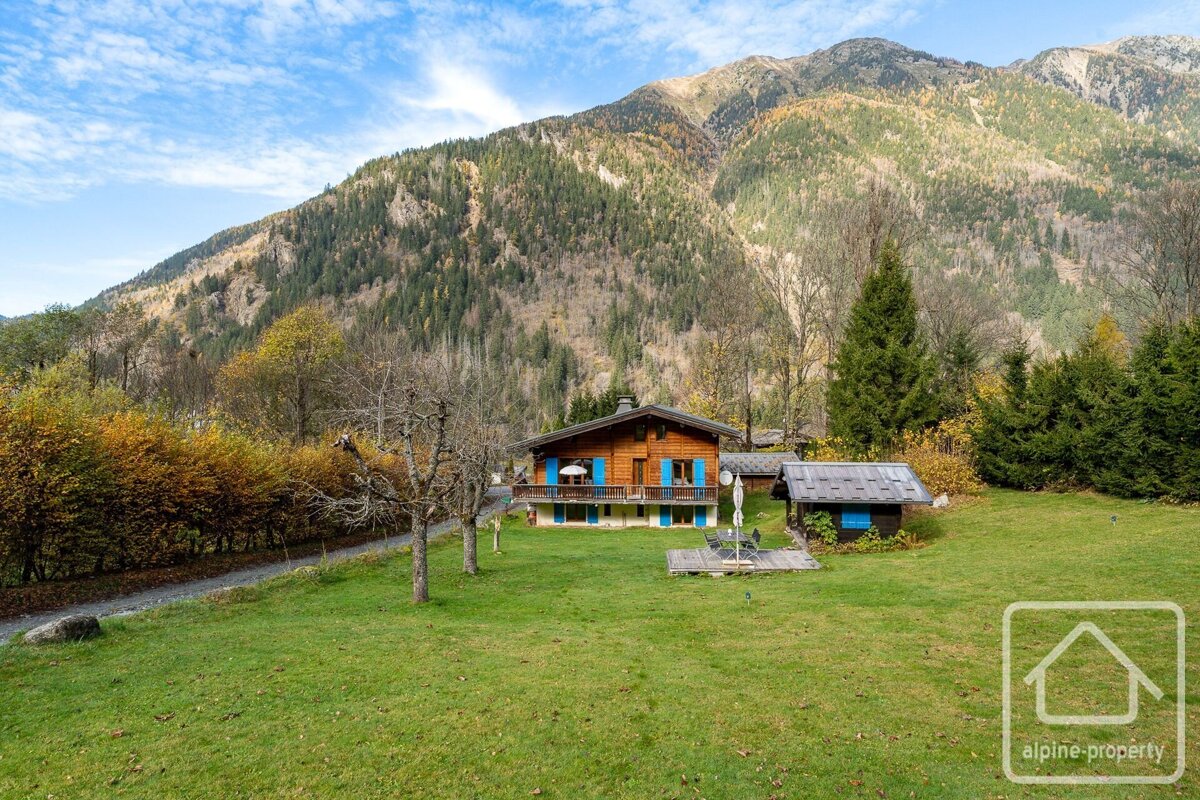 A wooden chalet with blue shutters stands on a green lawn, backed by a large, forested mountain under a cloudy sky. Autumn colors are visible.