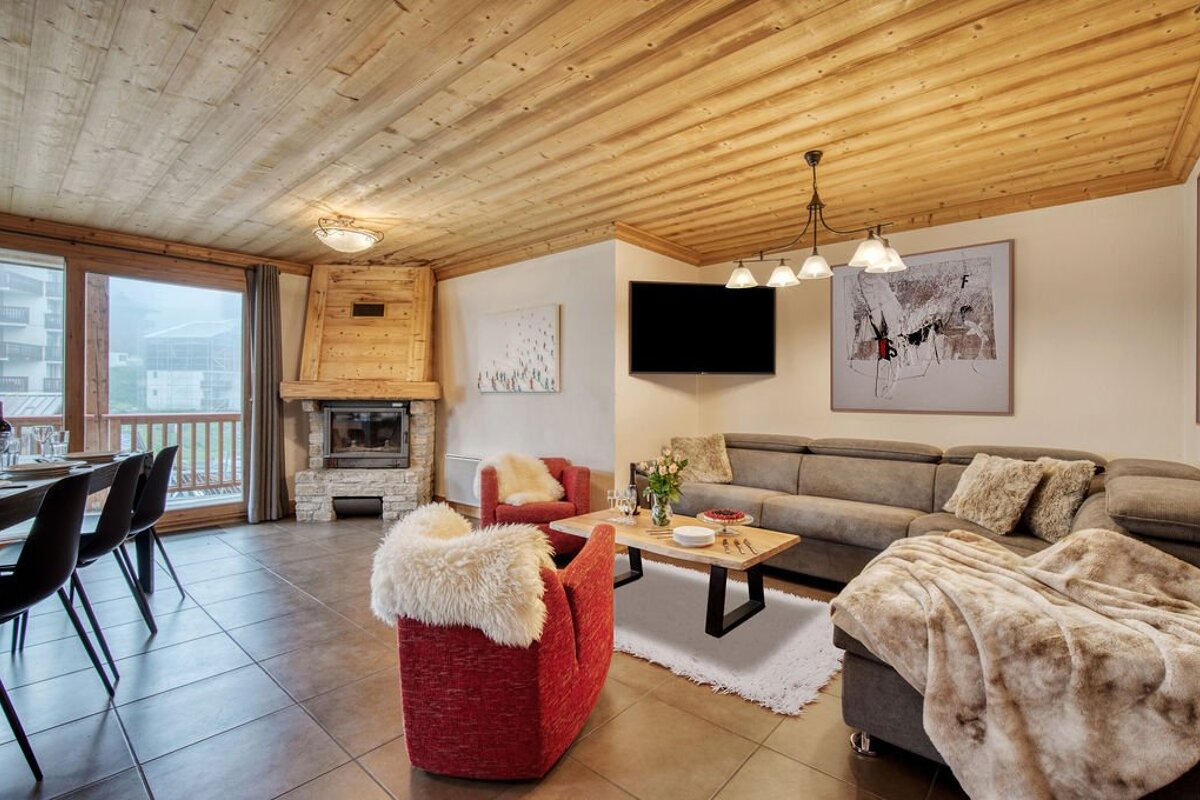 Cozy chalet living room with a wooden ceiling, grey sectional, red armchairs with white fur throws, coffee table, and corner fireplace.