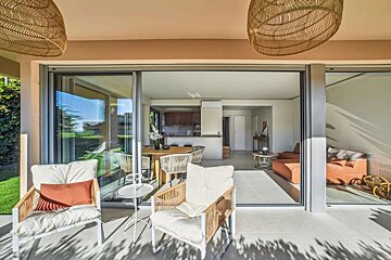A sunny patio with woven chairs overlooks a modern open-plan living space with a dining table, kitchen, and lounge, all seen through glass doors.