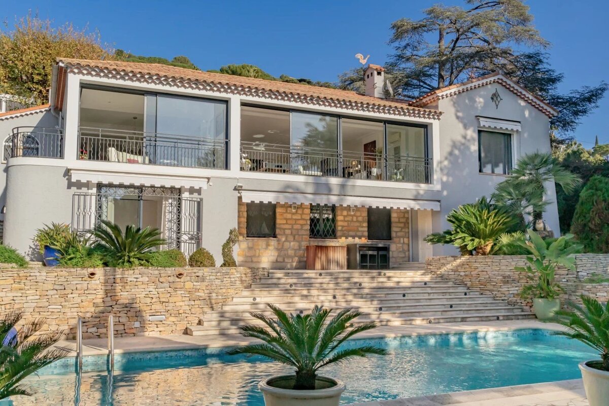 A luxurious two-story villa featuring a swimming pool, stone steps, and lush palm trees under a clear blue sky.