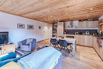 Open-plan living room and kitchen featuring wooden ceilings, light wood cabinets, and a dining table with chairs.