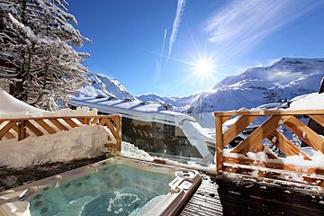 A bubbling hot tub on a snowy deck offers stunning views of sun-drenched mountains under a vibrant blue sky with contrails.