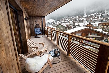 A cozy wooden chalet balcony with lounge chairs, overlooking a snowy alpine village nestled in foggy mountains.