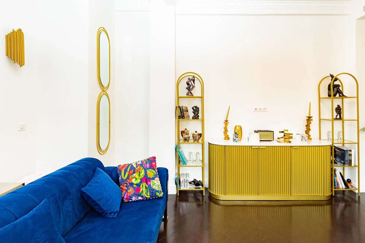 Stylish room featuring a royal blue sofa, gold fluted cabinet, arched shelves, and decorative mirrors on white walls with dark floors.