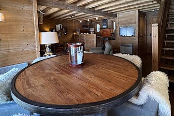 Cozy, rustic wooden chalet interior with a round table, exposed beams, and a view of the kitchen, living area, and stairs.