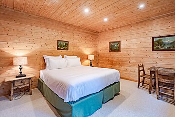 A cozy, wood-paneled bedroom with a white bed, two lamps on rustic nightstands, landscape art, and a small wooden table with chairs.