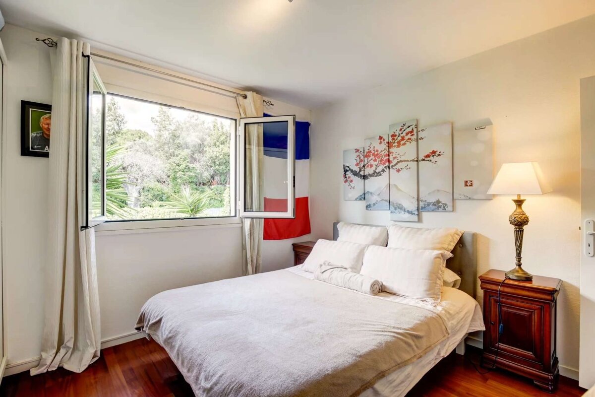 A bright bedroom with a large window overlooking greenery, a bed with white linens, cherry blossom art, and a French flag visible.