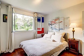 A bright bedroom with a large window overlooking greenery, a bed with white linens, cherry blossom art, and a French flag visible.