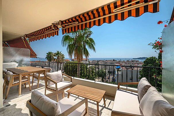 A bright balcony featuring orange striped awnings, wooden outdoor furniture, lush palm trees, and a scenic view of the city and sea.