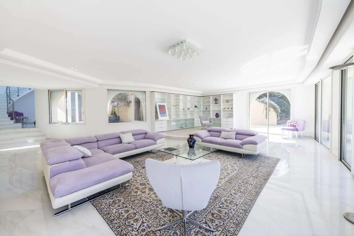 A bright, spacious living room featuring lavender sofas, a patterned rug, glass coffee table, built-in shelves, and large windows.