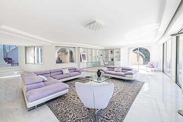A bright, spacious living room featuring lavender sofas, a patterned rug, glass coffee table, built-in shelves, and large windows.