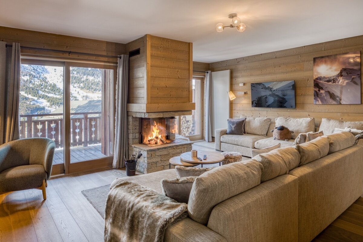 Cozy wooden chalet living room with a warm stone fireplace, comfortable sofas, and snowy mountain views through the glass doors.