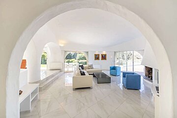 Spacious, modern white living room with arched walls, tiled floor, cream sectional sofa, blue armchairs, and large windows opening to a garden.