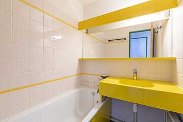 A bathroom with white tiled walls accented by yellow stripes. It has a white bathtub, a bright yellow sink, and a large mirror.