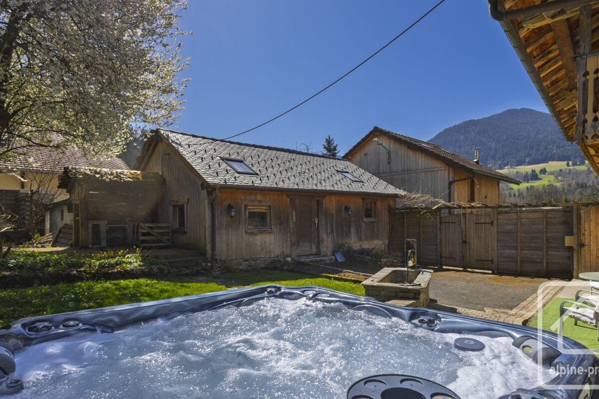 A bubbling hot tub in a sunny yard with a rustic wooden cabin and distant mountains under a clear blue sky.