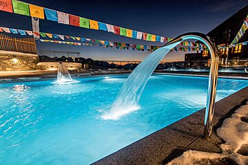 An illuminated outdoor pool at dusk with waterfalls, surrounded by snow and colorful prayer flags, against a scenic mountain backdrop.