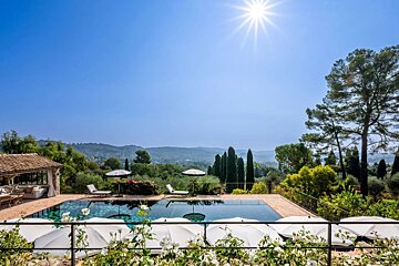 Sunny view of a luxury villa pool surrounded by lush greenery and a bright blue sky.
