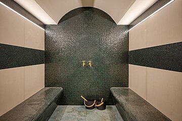 Tiled steam room with dark green mosaic walls, two benches, brass faucets, strip lighting, and two buckets on the floor.