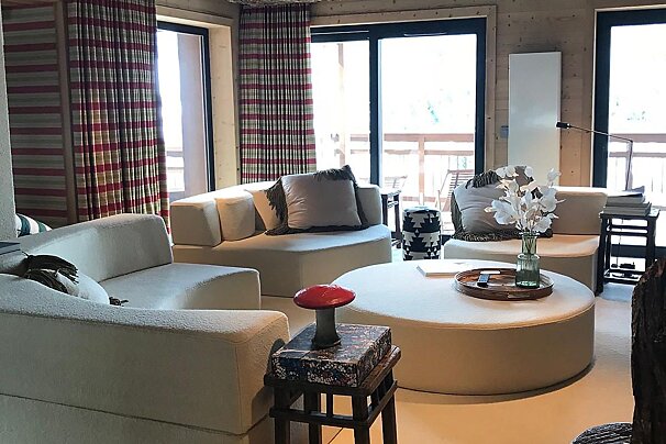 A cozy living room with cream modular sofas, a round coffee table, and plaid curtains, offering a view of the outdoors through large windows.