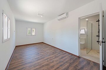 An empty, brightly lit room with white walls, wood floor, windows, an AC unit, and an open doorway to an en-suite bathroom with a toilet and shower.