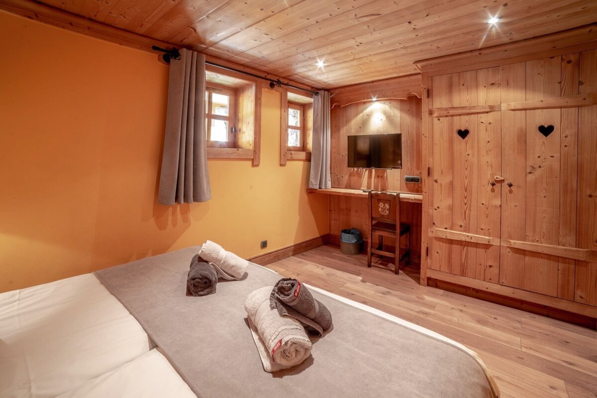 A rustic, cozy bedroom with a wood-paneled ceiling, orange accent wall, bed with towels, windows, a TV, and a charming wooden wardrobe with heart cutouts.