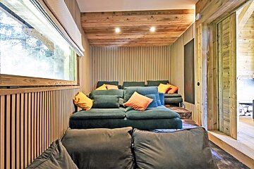 Cozy, wood-paneled home cinema with tiered dark green sofas, bright orange/yellow pillows, and a window. Rustic chalet interior.