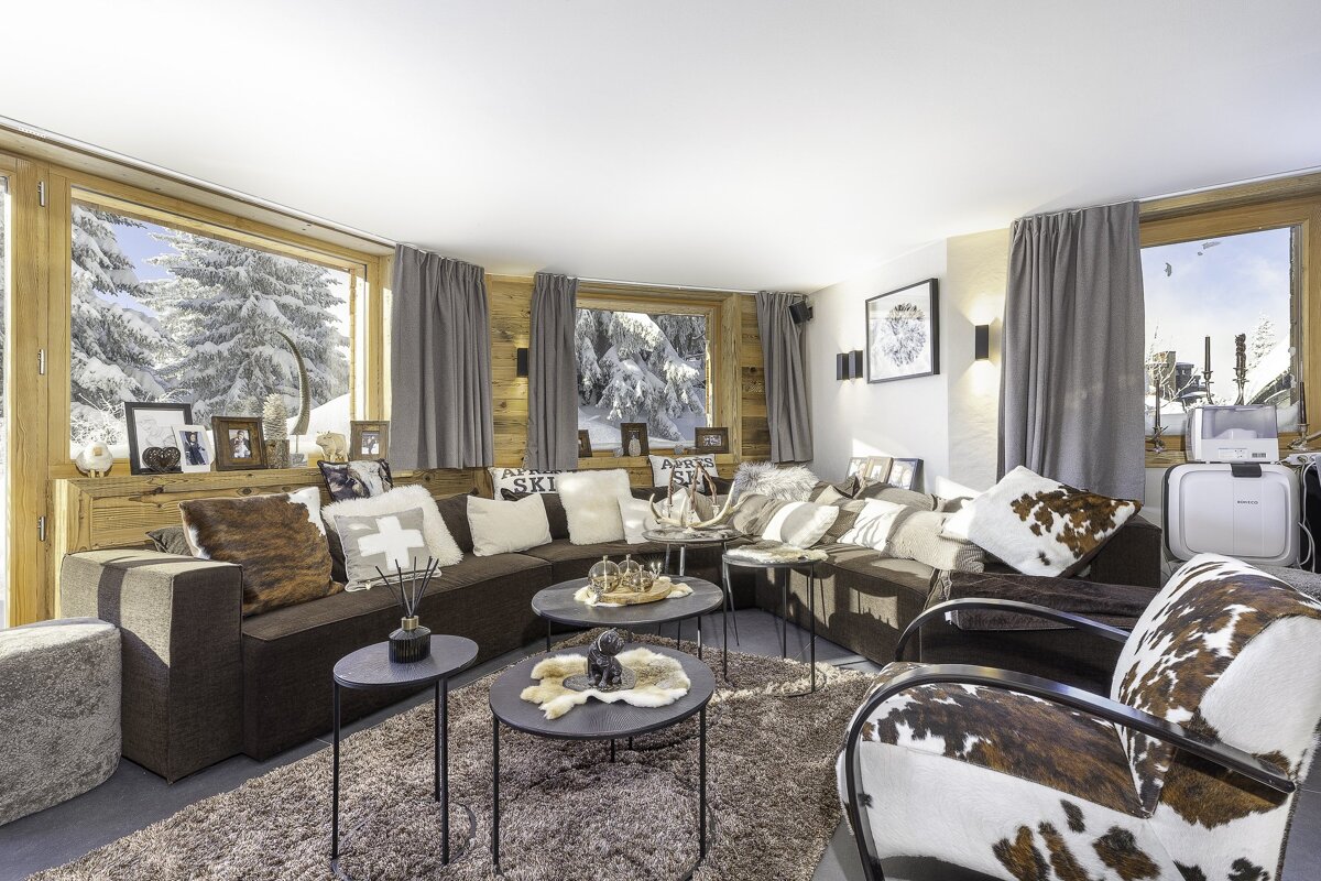 A cozy, rustic chalet living room with a large brown sofa, cowhide accents, and snowy winter views through the windows.