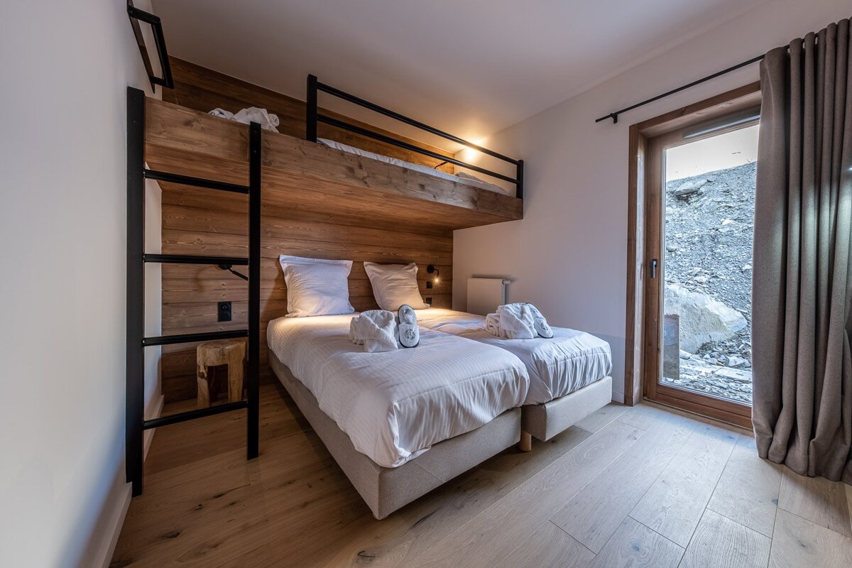 Modern bedroom featuring a built-in bunk bed above two single beds, wood paneling, light wooden floor, and a door/window with a rocky outdoor view.