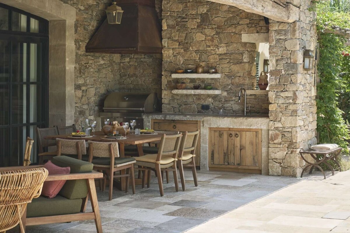 A rustic outdoor kitchen and dining area with stone walls, a built-in grill, sink, and a dining table set for guests on a stone patio.
