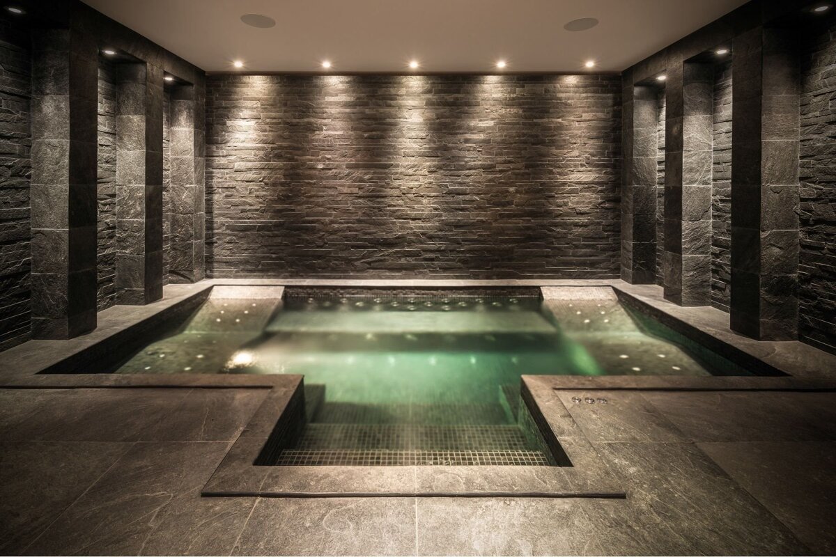 Luxurious indoor spa with dark stone walls, a turquoise water hot tub, and recessed lighting creating a serene, modern ambiance.