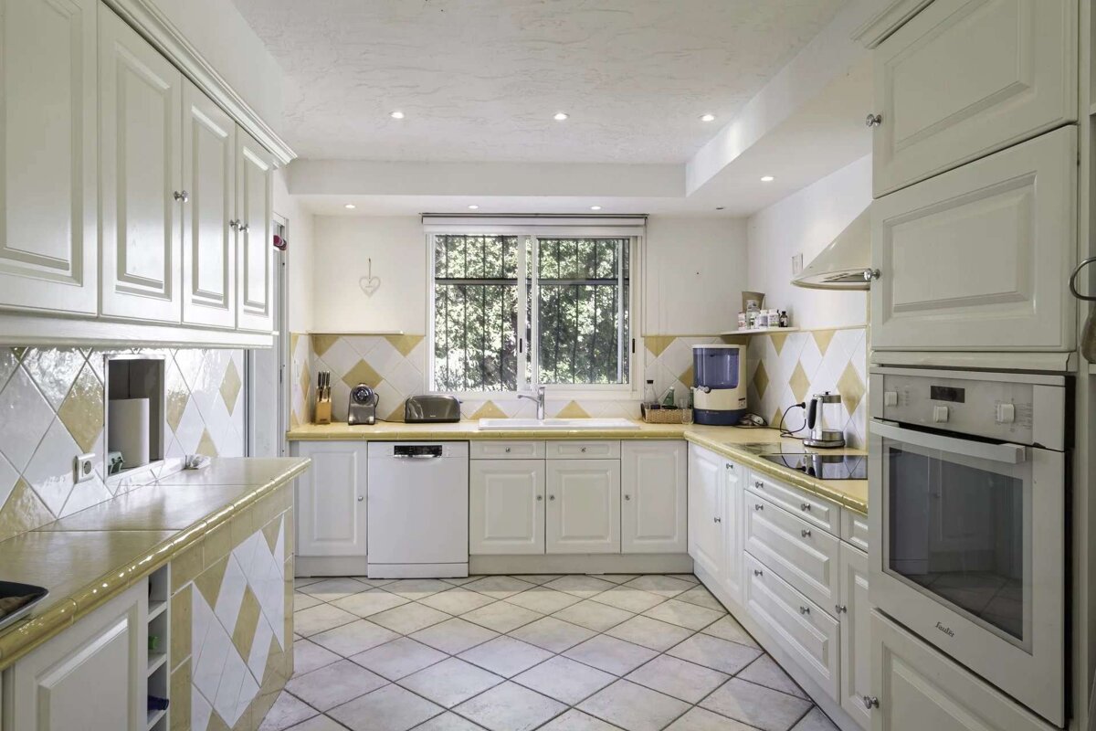 A bright kitchen with white cabinets, yellow countertops, and diamond-patterned tiles on the floor and backsplash. A window overlooks greenery.