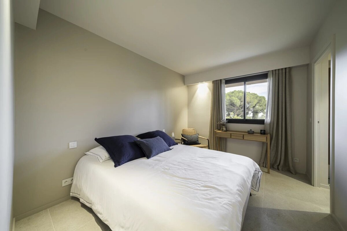 Minimalist bedroom with a large bed, neutral walls, and a window displaying trees outside.
