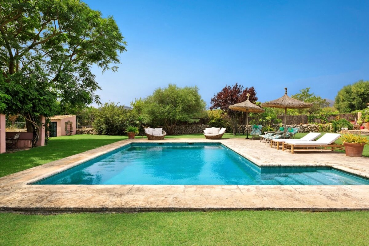 Sunny backyard with a bright blue pool, green lawn, stone patio, lounge chairs, straw umbrellas, and lush trees under a clear sky.