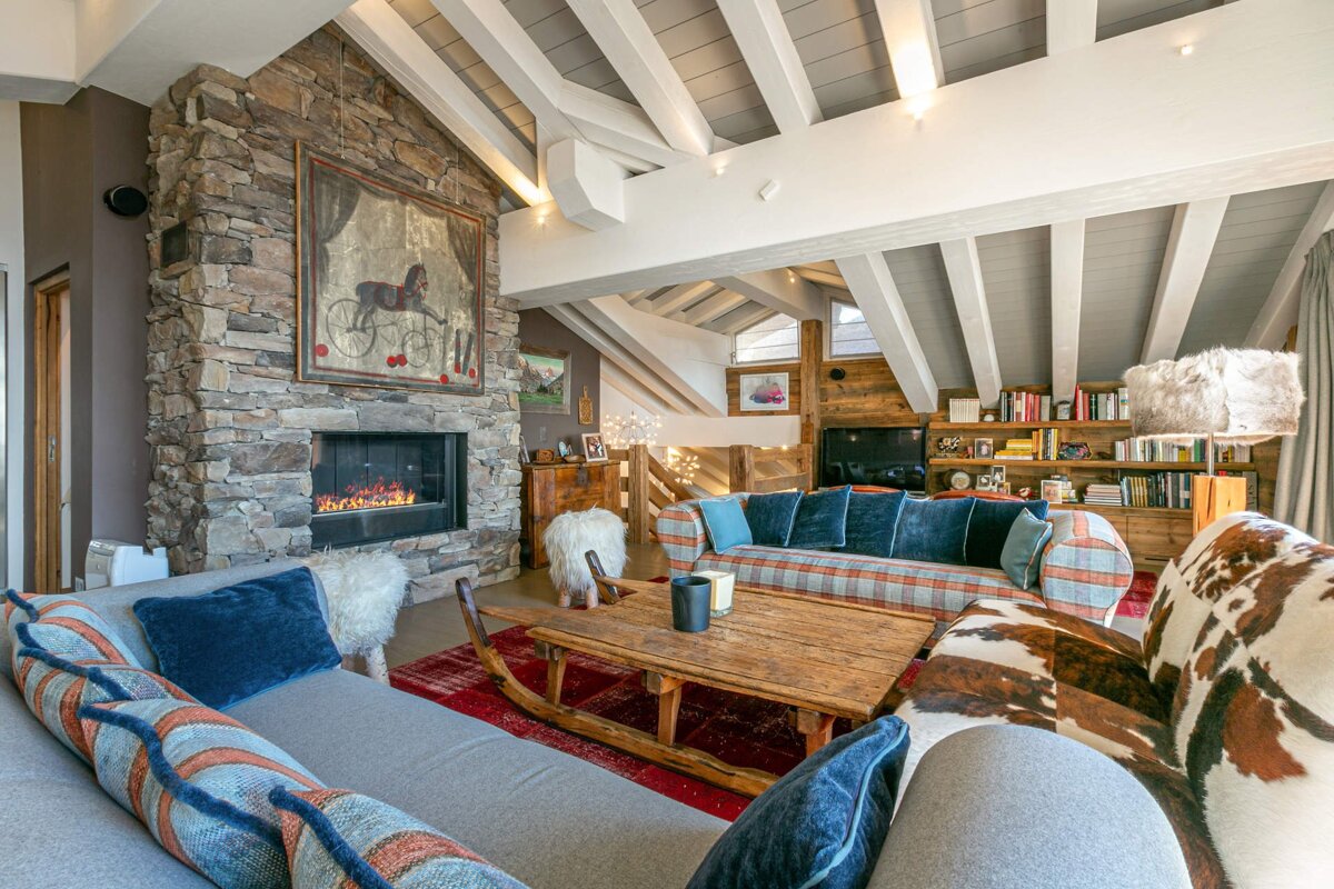 Cozy chalet living room featuring a stone fireplace, vaulted beamed ceiling, sled coffee table, and eclectic sofas.