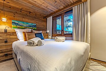 Cozy alpine-style bedroom with wood-paneled walls and ceiling, featuring a bed with white linens, colorful pillows, and a window view.