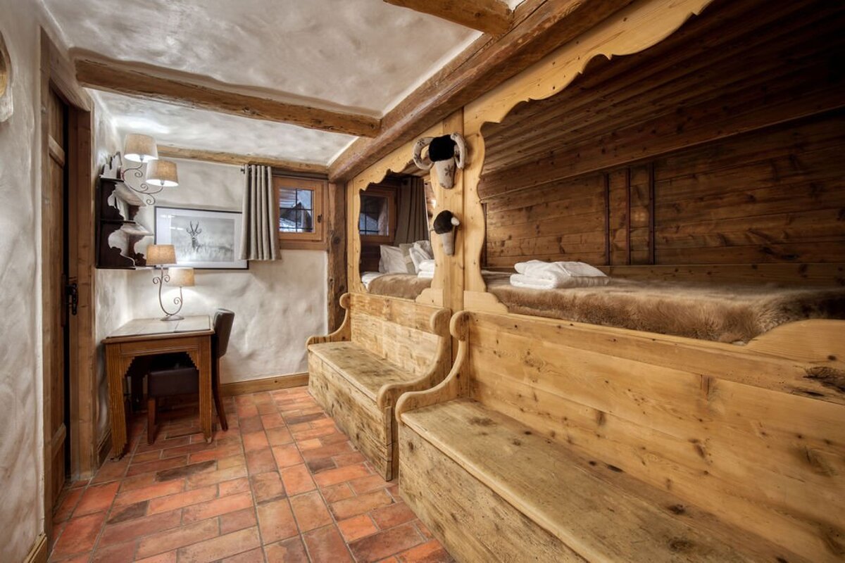 Cozy rustic cabin room with built-in wooden beds and benches, plaster walls, a small corner desk, and terracotta tile flooring.