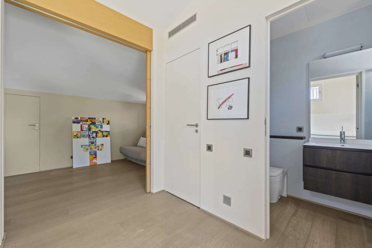 A modern hallway with light wood floors connects to an open attic-style room and a minimalist bathroom. White walls display framed artwork.