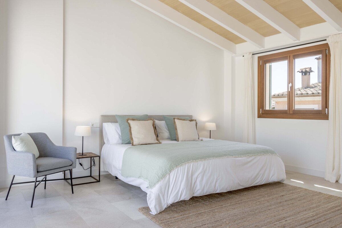 A bright, minimalist bedroom featuring a bed with light blue accents, a grey armchair, a window with a rooftop view, and a sloped ceiling with wooden beams.