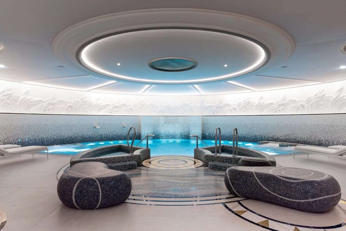 A luxurious, circular indoor spa with a blue pool, two whirlpools, mosaic walls, and elegant ambient lighting beneath a domed ceiling.