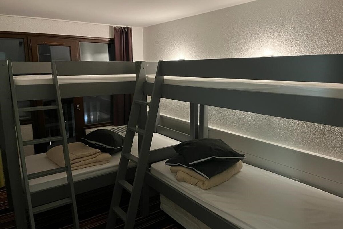 Grey bunk beds with white mattresses, dark pillows, and light blankets. Ambient wall lighting illuminates a textured wall, with a window visible.