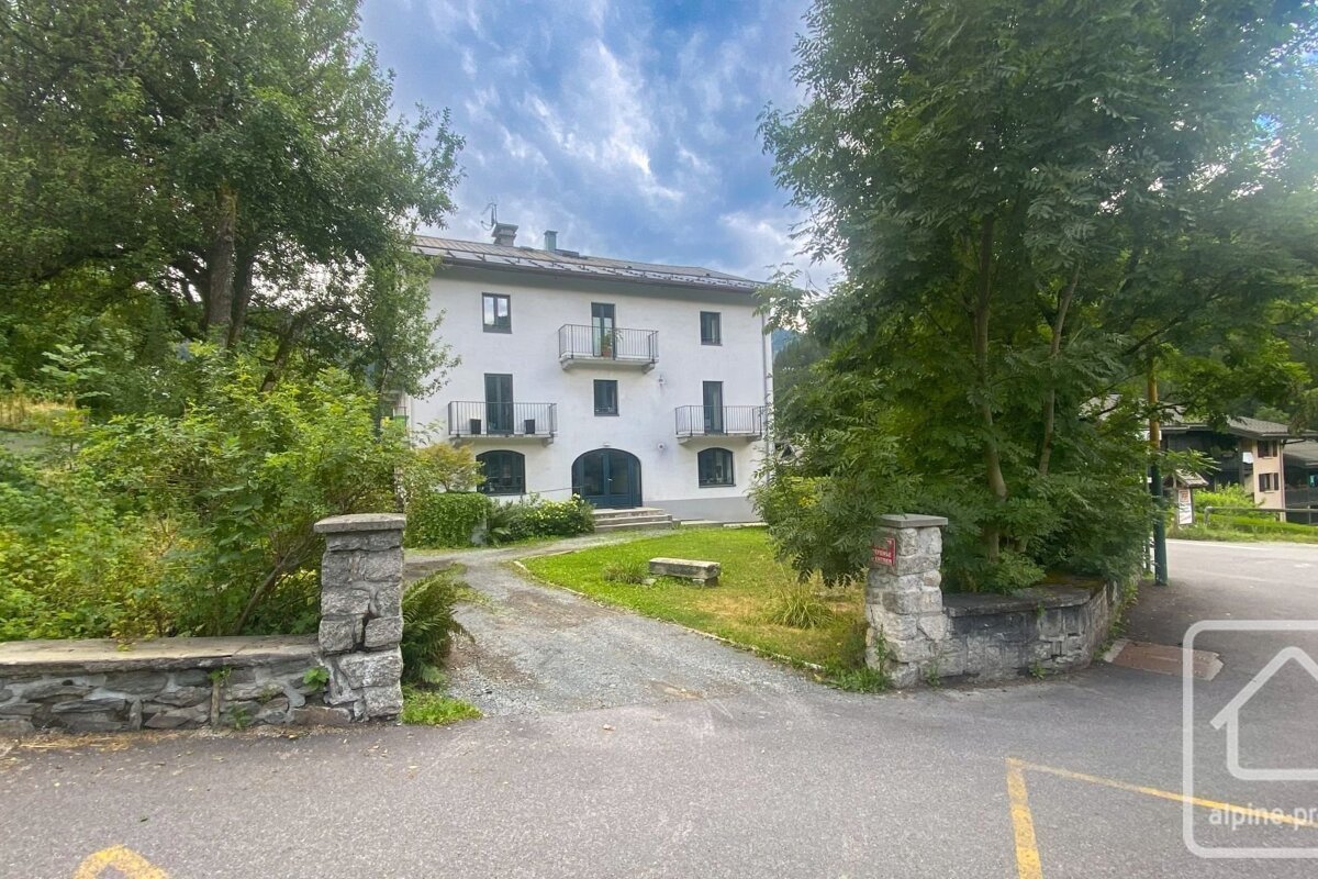 A white building with balconies is nestled among green trees, featuring a gravel driveway and stone pillars, under a cloudy sky.