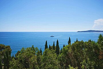 Expansive blue sea under a clear sky with a distant yacht. Lush green trees frame the foreground, leading to a far-off coastline.