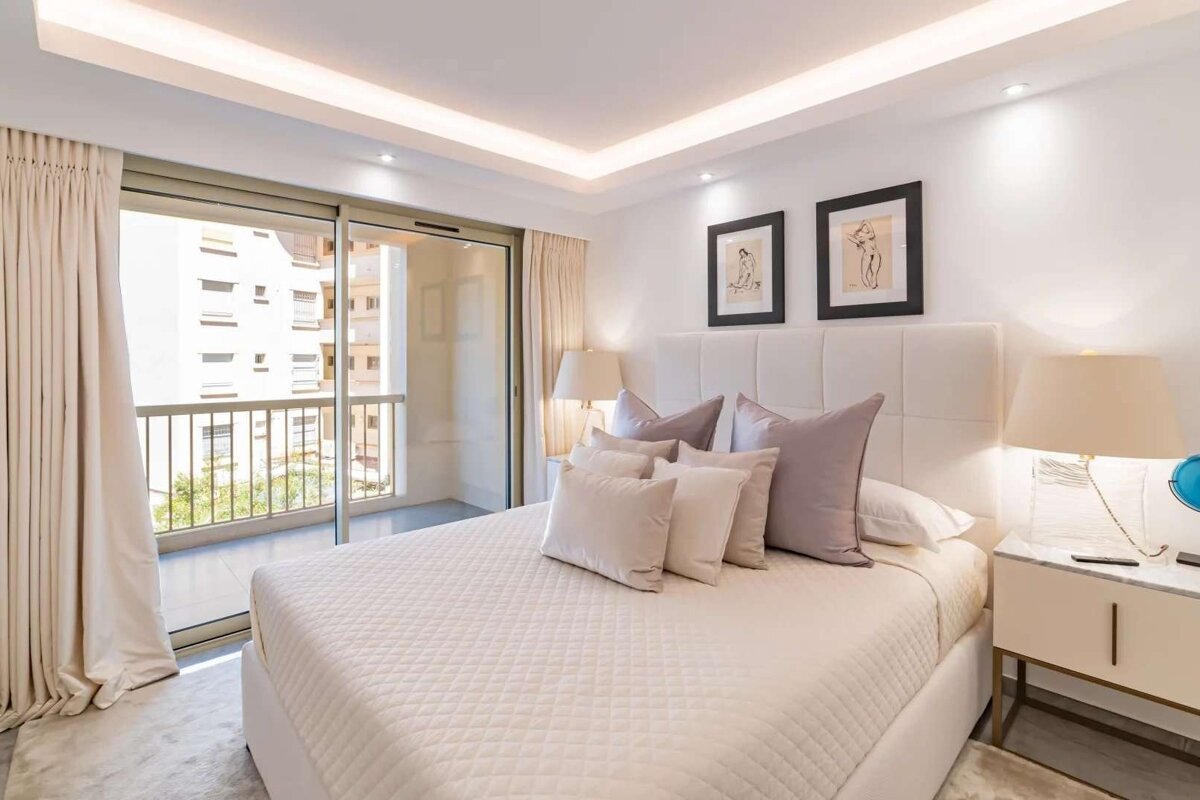 Bright, modern bedroom in neutral tones with a large bed, artwork, and a sliding glass door opening to a balcony view.